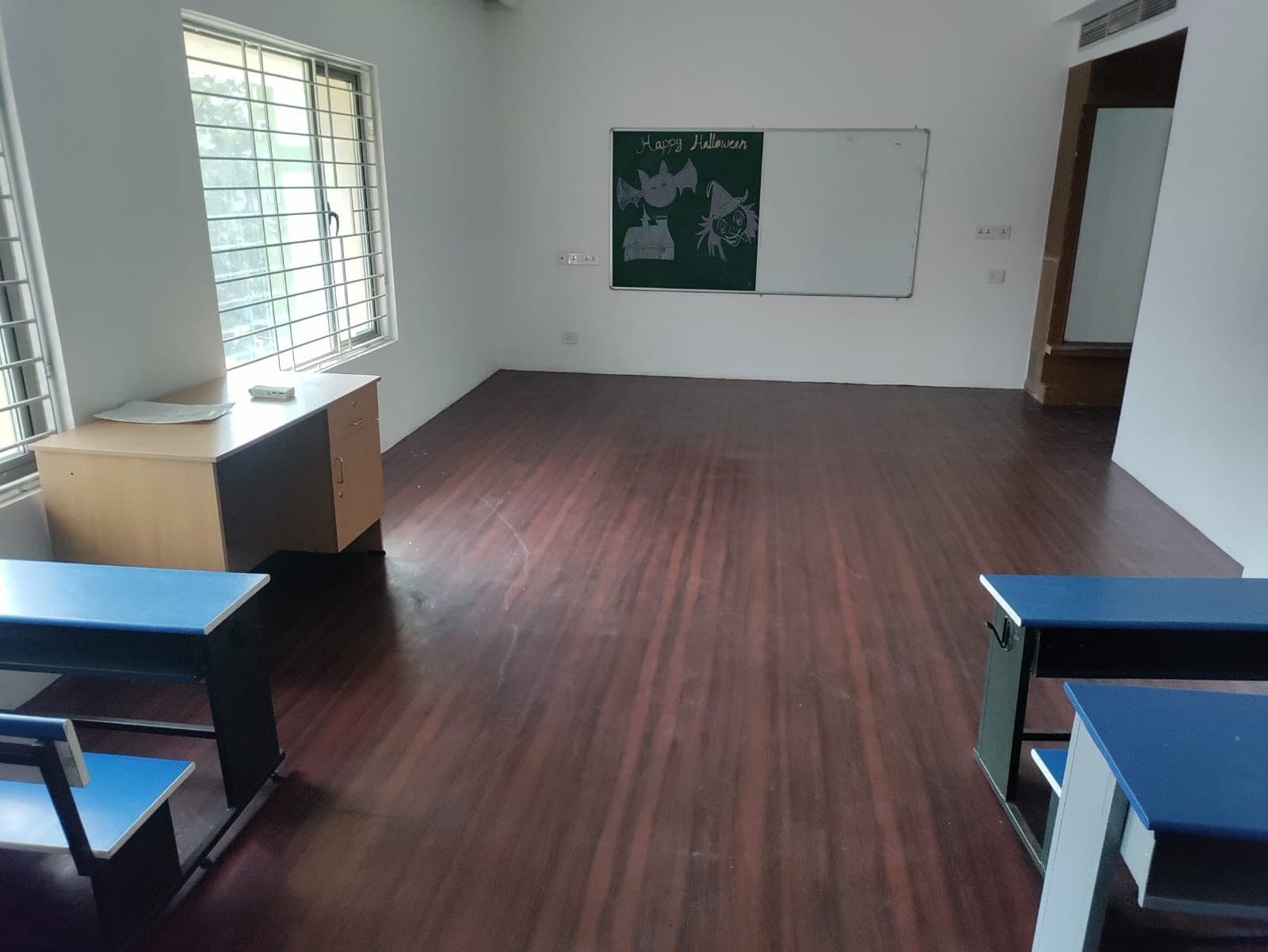 Completed classroom in Chennai with timber-effect flooring, a combined chalk and marker board on the end wall, a teacher's desk by the window and pupils' desks in the foreground.