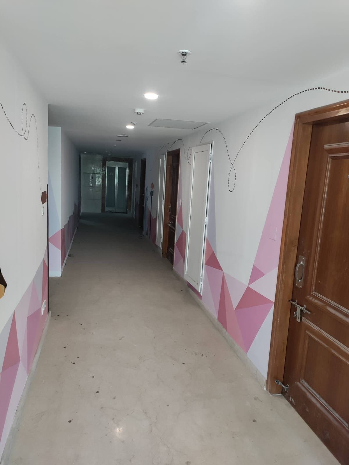 The same building on another floor, the corridor finished in a pink and mauve geometric scheme, with the lift lobby at the far end and sprinkler heads in the ceiling.