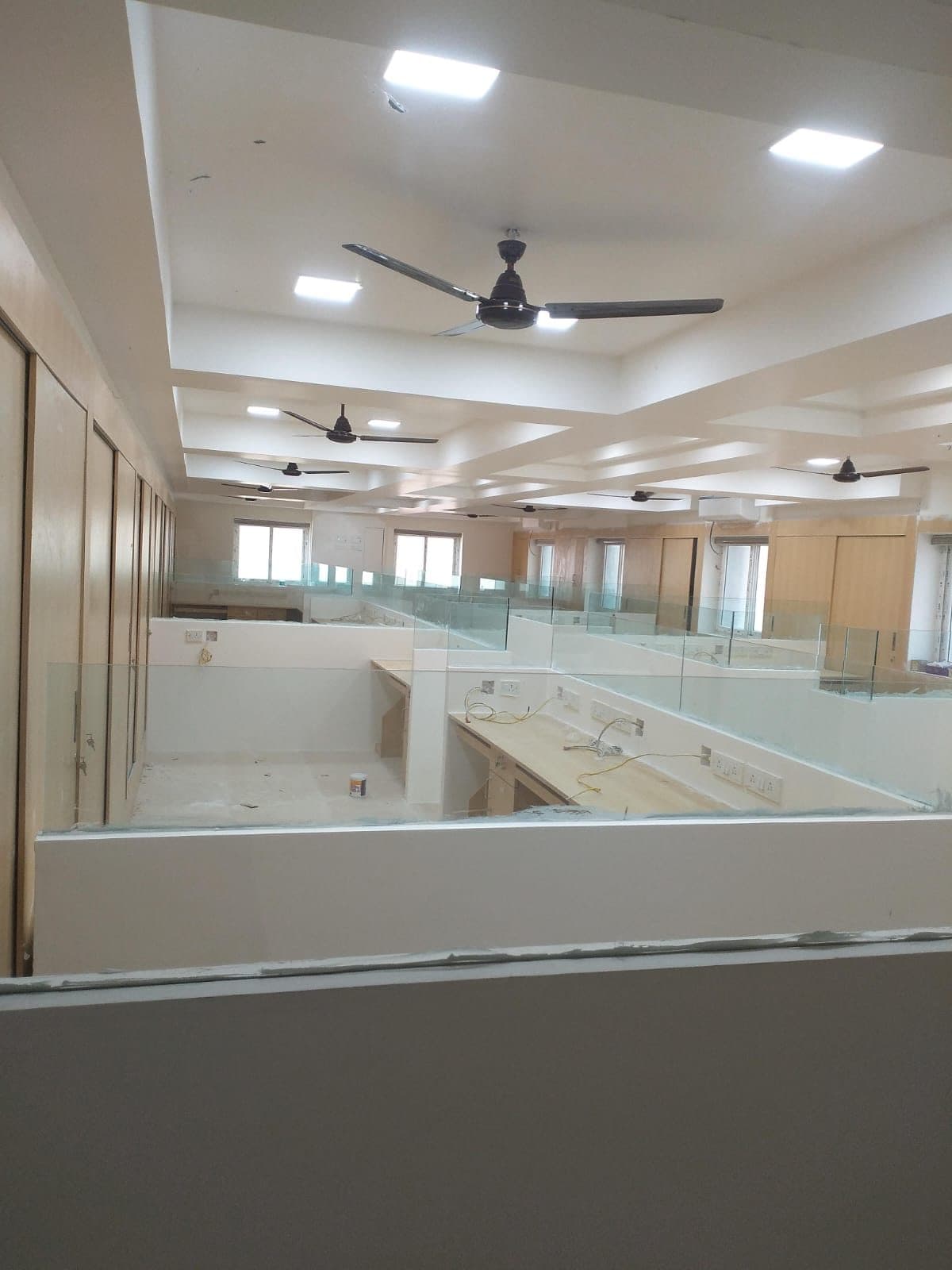 Open office floor with glass-partitioned workstations, built-in desking and a stepped false ceiling carrying recessed lights and ceiling fans.