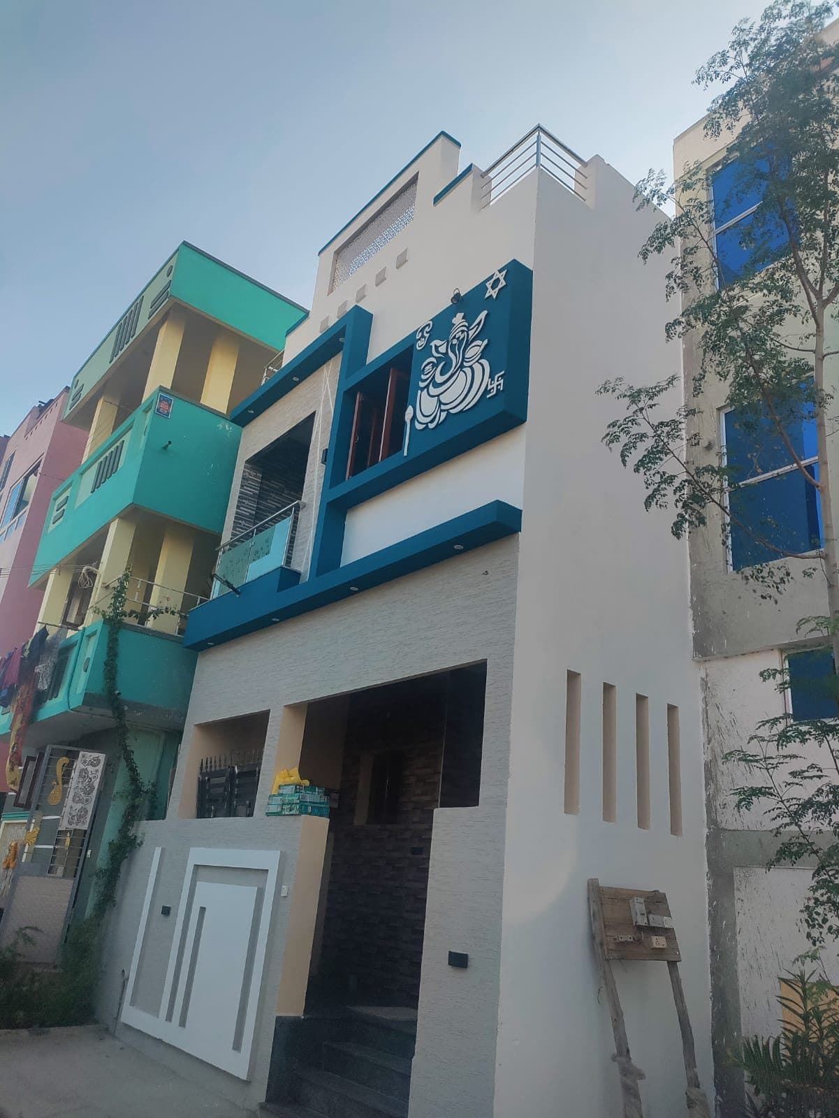 Completed narrow-plot two-storey house in Chennai, white render with teal banding, split-face stone cladding, a cast relief panel above the first-floor window and an etched glass balcony railing.