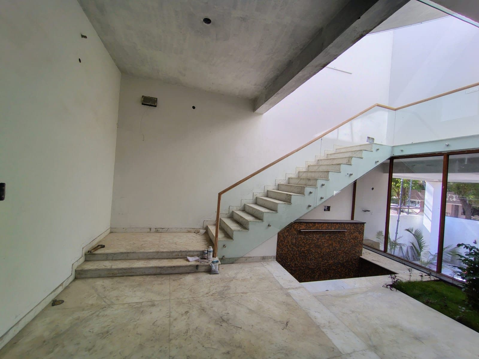 Double-height entrance hall in Chennai with a marble-clad stair, glass balustrade on a timber handrail, marble floor, mosaic-faced planter and full-height glazing to an internal courtyard.