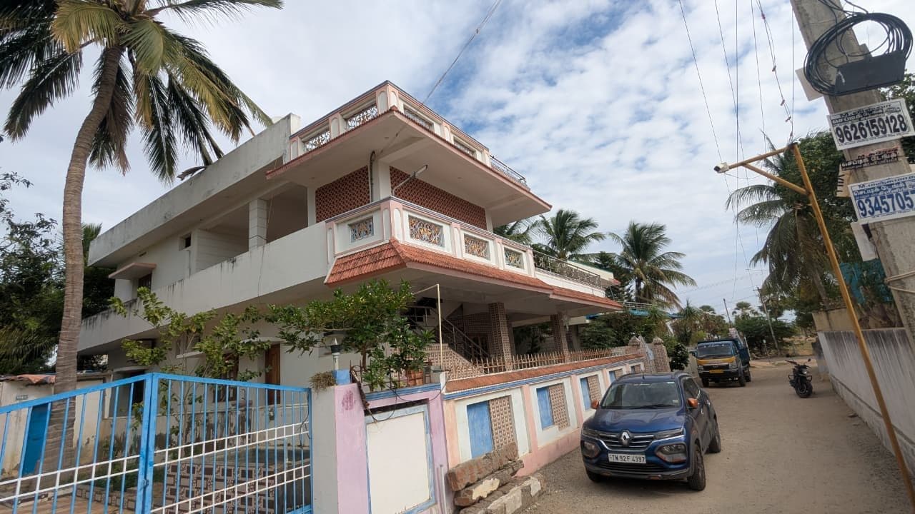 Corner view of a completed two-storey residence with clay-tiled sunshades, perforated jaali balcony panels and an external stair, behind a brick-clad compound wall.
