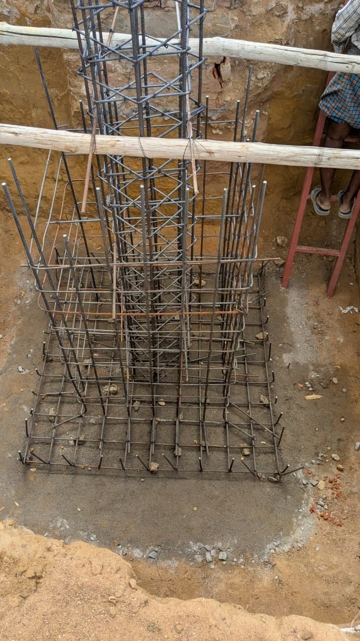 Isolated footing in open excavation: two-way reinforcement mesh tied over a plain cement concrete bed, with a plumbed column starter cage rising from the centre and timber barricade rails across the pit.