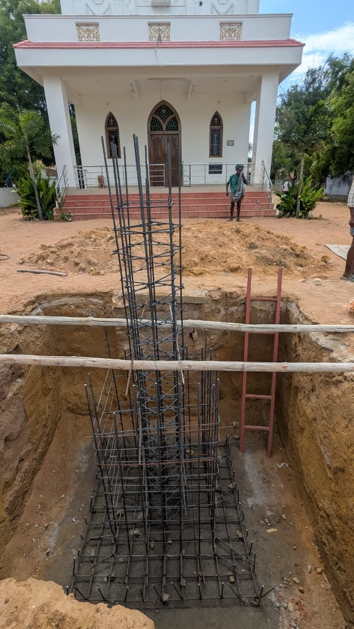 Column foundation under construction in the compound of a church, showing the excavation, reinforcement cage, spoil heap and an access ladder into the pit.
